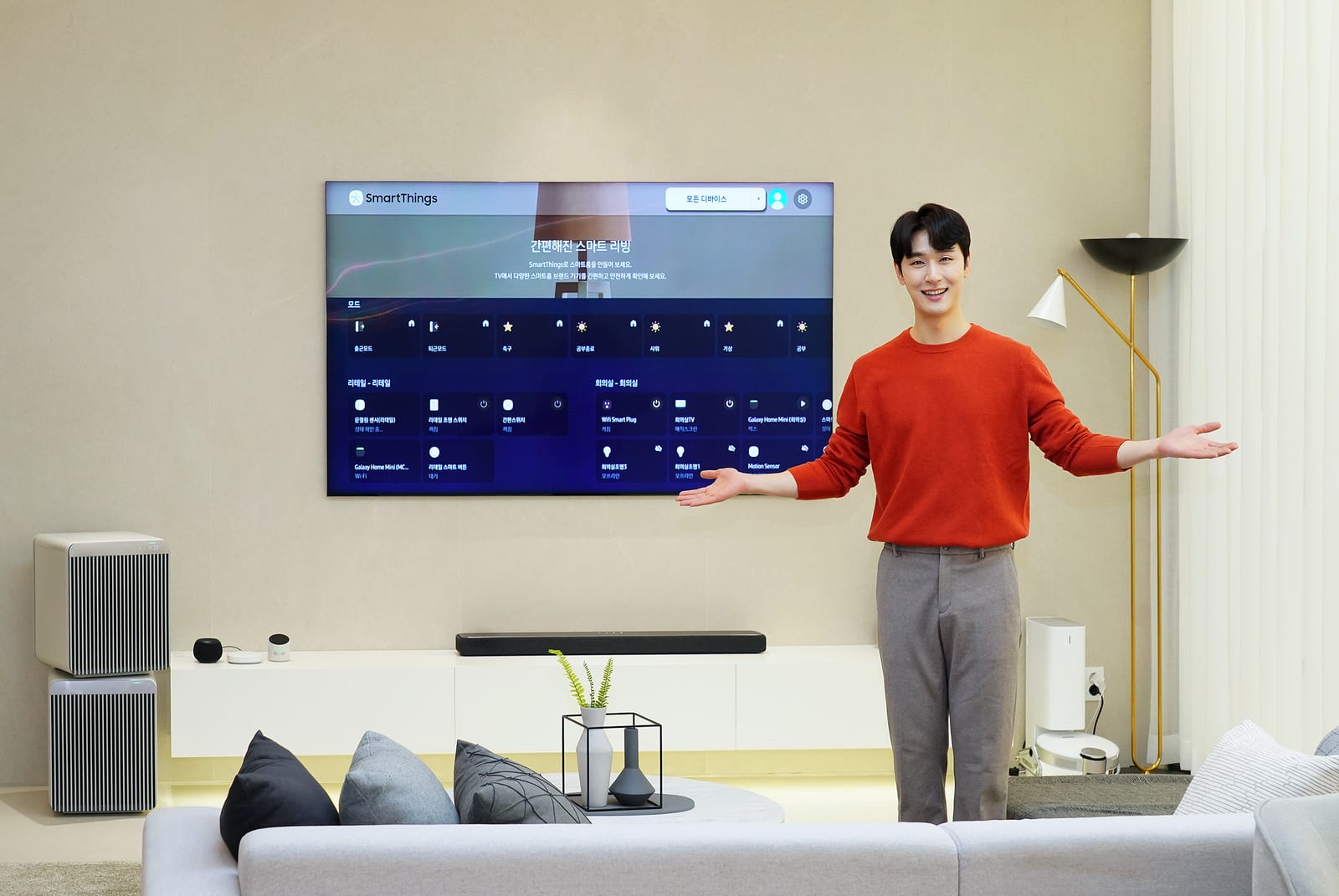 스마트 아파트 거실. 벽걸이 TV에 SmartThings 화면이 켜져 있고 모델이 양팔을 펼쳐 소개하고 있다.